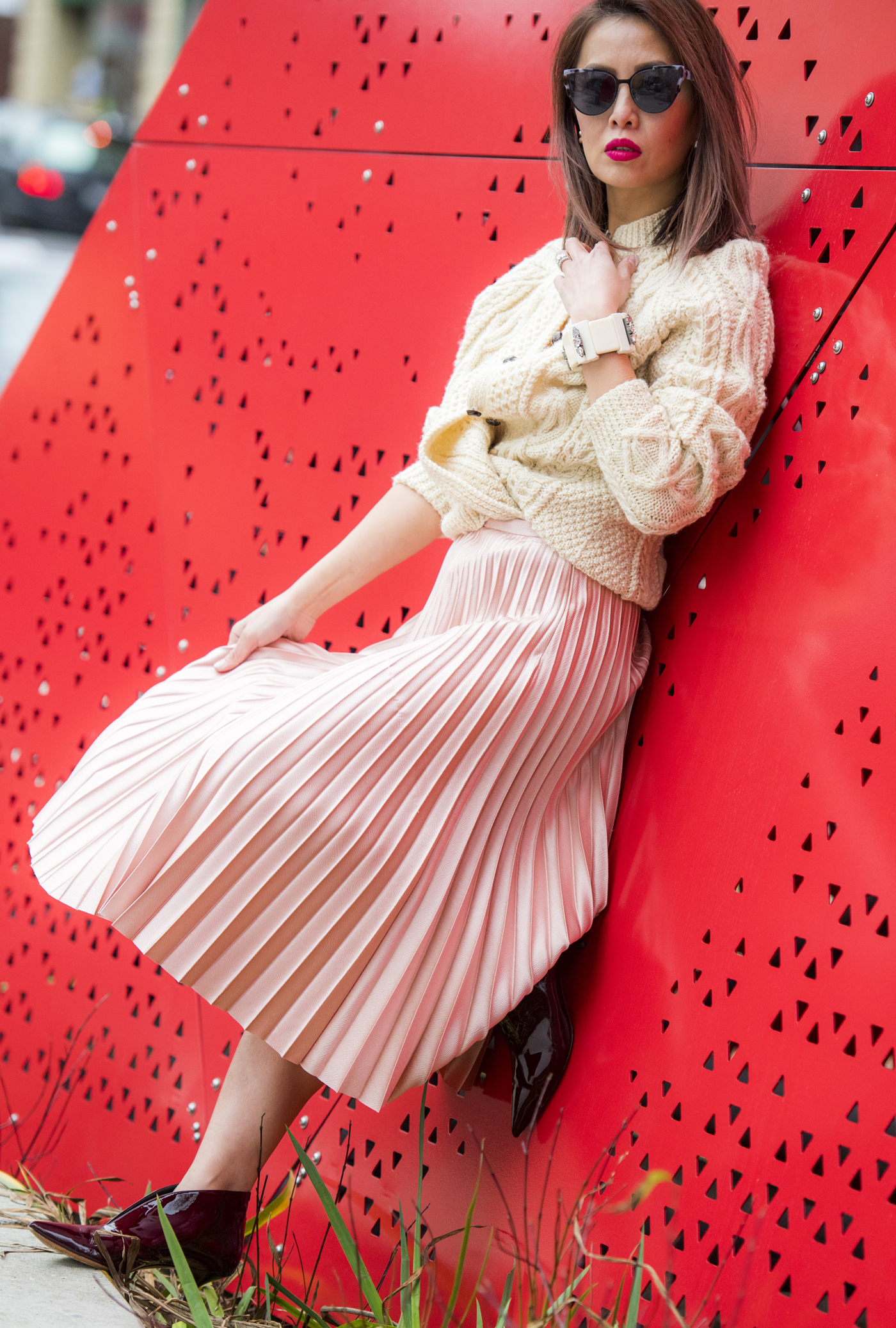 Elisa-Pink-Pleated-Skirt-White-Cable-Cardigan-Leaning-on-Red-Sculpture.jpg